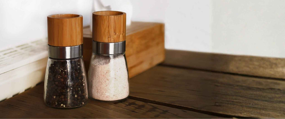 Custom salt and pepper mill set with wooden tops and clear glass bodies on a wooden kitchen countertop.