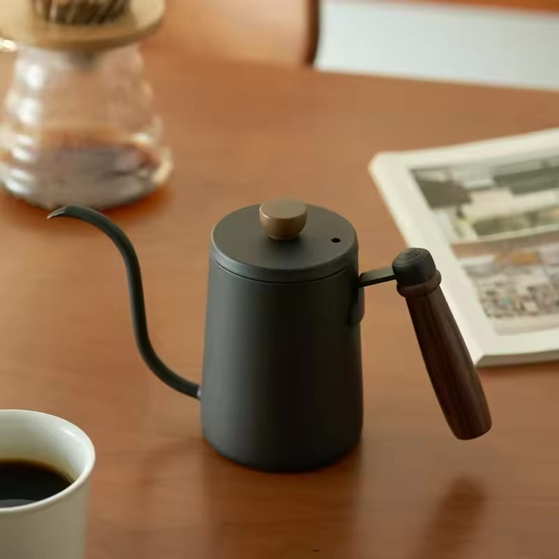 Matte black gooseneck kettle with a wood handle beside a cup of coffee.